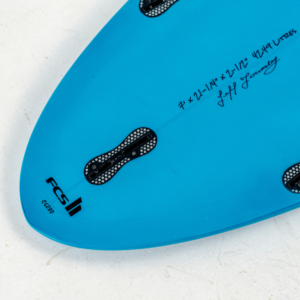 Close-up of a bright blue FCS Li egg surfboard showing the underside with black fin hardware, handwritten signatures, and the model number C0010 on a white textured surface.