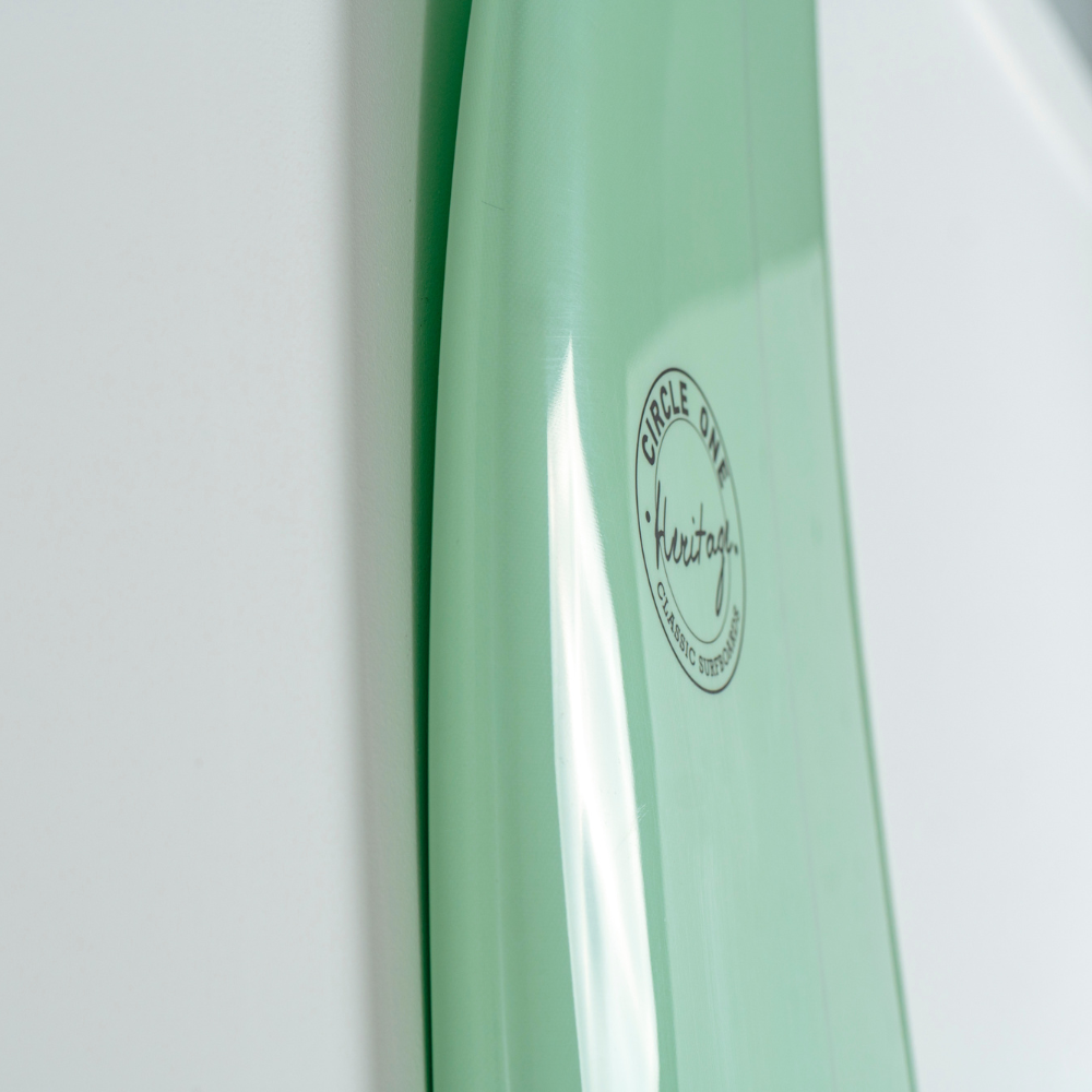 Close-up of a mint green egg surfboard showing the side with a circular black logo that reads 'Circle Gains Kehehau'.
