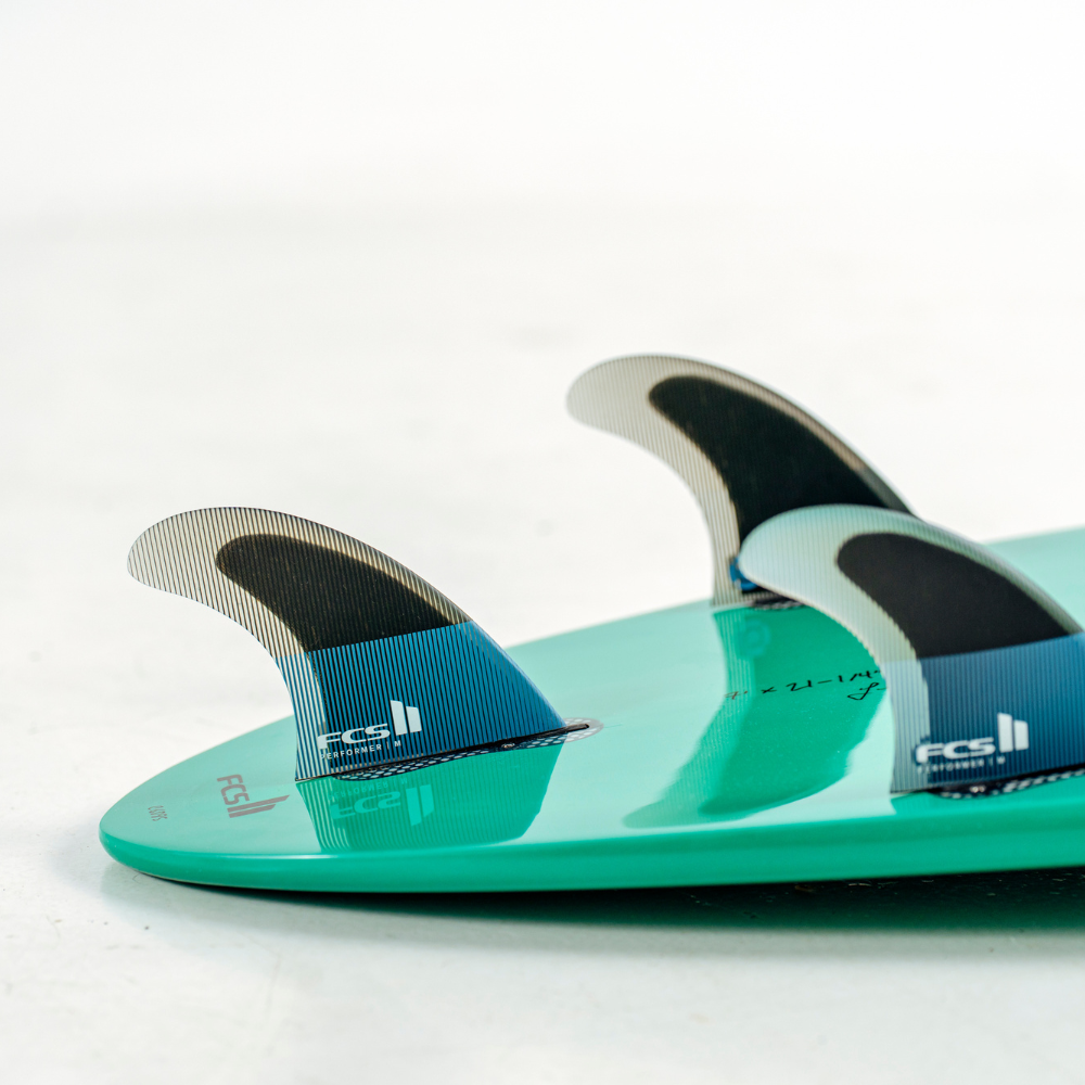 Close-up of a surfboard tail with three FCS Ultimate 11 fins, featuring a bright teal-green deck and blue and black fin blades on a white background.
